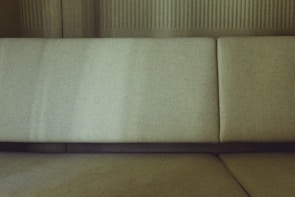 A close-up view of a minimalist sofa with a textured fabric finish. The upholstery features a subtle olive green tone, set against a backdrop of matching tones. The fabric appears softly lit, highlighting its natural texture.