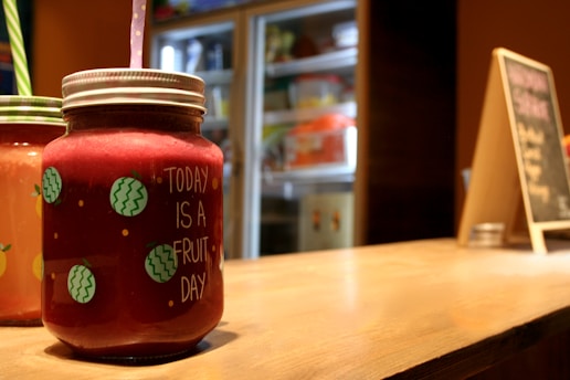 A close-up of a jar filled with a thick red beverage, labeled 'Today is a fruit day' with decorative illustrations of fruits. The jar has a lid and a straw sticking out of it. In the background, there's a blurred view of a refrigerator and a chalkboard menu, both out of focus. The overall setting appears to be a small café or juice bar.