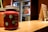 A close-up of a jar filled with a thick red beverage, labeled 'Today is a fruit day' with decorative illustrations of fruits. The jar has a lid and a straw sticking out of it. In the background, there's a blurred view of a refrigerator and a chalkboard menu, both out of focus. The overall setting appears to be a small café or juice bar.