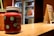 A close-up of a jar filled with a thick red beverage, labeled 'Today is a fruit day' with decorative illustrations of fruits. The jar has a lid and a straw sticking out of it. In the background, there's a blurred view of a refrigerator and a chalkboard menu, both out of focus. The overall setting appears to be a small café or juice bar.