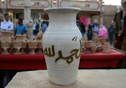 A traditional ceramic vase with Arabic calligraphy is prominently displayed on a ledge. The background shows a busy market setting with people and various pottery items on display.