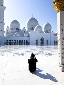 A person sits on a polished white marble floor, capturing a photo of a grand mosque with multiple large domes and intricate architectural details. The mosque is bathed in sunlight, and its pristine white surface contrasts with the clear blue sky. Floral designs can be seen on a column to the right, and the expansive courtyard is decorated with subtle artistic patterns.