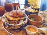 A sesame seed hamburger bun with a juicy patty, melted cheese, and lettuce is the main focus, placed in a lined basket. Accompanying the burger are small cups of dipping sauces, and a box of golden French fries. Tall glasses filled with iced beverages can be seen in the background.
