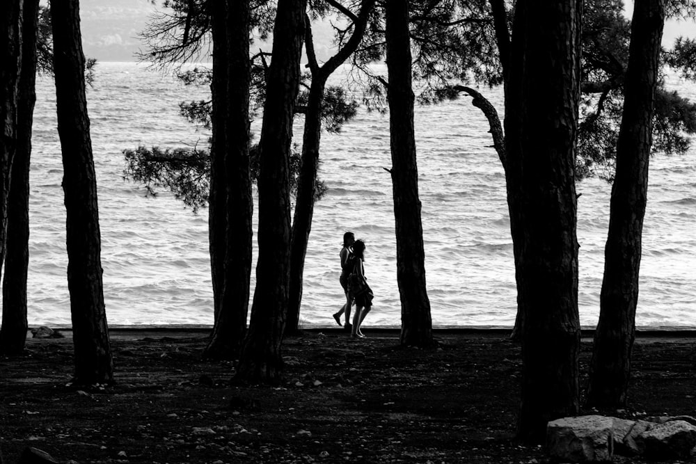 grayscale photo of two persons walking beside body of water surrounded with trees
