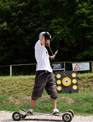 A person is skillfully balancing on an all-terrain skateboard while drawing a bow and arrow in an archery range. The background features a lush green forest and a target board with yellow circles.
