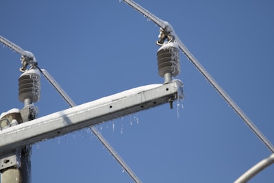 A section of an electrical power line covered in ice and snow against a clear blue sky. The cables and insulators are thickly layered with ice, indicating cold weather conditions.
