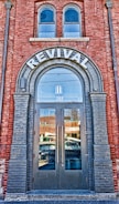 A brick building facade featuring a large arched doorway labeled 'REVIVAL' at the top. The doorway has double glass doors with tall vertical handles. Above the door, there are two arched windows. The reflection in the glass doors shows a busy street with passing vehicles and nearby buildings. The brickwork around the entrance is accented with darker bricks framing the arch and the doorway.