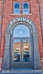 A brick building facade featuring a large arched doorway labeled 'REVIVAL' at the top. The doorway has double glass doors with tall vertical handles. Above the door, there are two arched windows. The reflection in the glass doors shows a busy street with passing vehicles and nearby buildings. The brickwork around the entrance is accented with darker bricks framing the arch and the doorway.