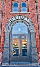 A brick building facade featuring a large arched doorway labeled 'REVIVAL' at the top. The doorway has double glass doors with tall vertical handles. Above the door, there are two arched windows. The reflection in the glass doors shows a busy street with passing vehicles and nearby buildings. The brickwork around the entrance is accented with darker bricks framing the arch and the doorway.