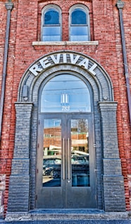 A brick building facade featuring a large arched doorway labeled 'REVIVAL' at the top. The doorway has double glass doors with tall vertical handles. Above the door, there are two arched windows. The reflection in the glass doors shows a busy street with passing vehicles and nearby buildings. The brickwork around the entrance is accented with darker bricks framing the arch and the doorway.