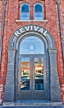 A brick building facade featuring a large arched doorway labeled 'REVIVAL' at the top. The doorway has double glass doors with tall vertical handles. Above the door, there are two arched windows. The reflection in the glass doors shows a busy street with passing vehicles and nearby buildings. The brickwork around the entrance is accented with darker bricks framing the arch and the doorway.