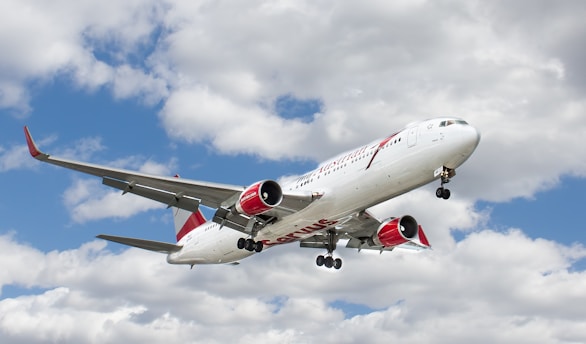 A large commercial airplane is seen flying in the sky with a backdrop of fluffy white clouds. The aircraft has a white body with red accents on the engines and tail.