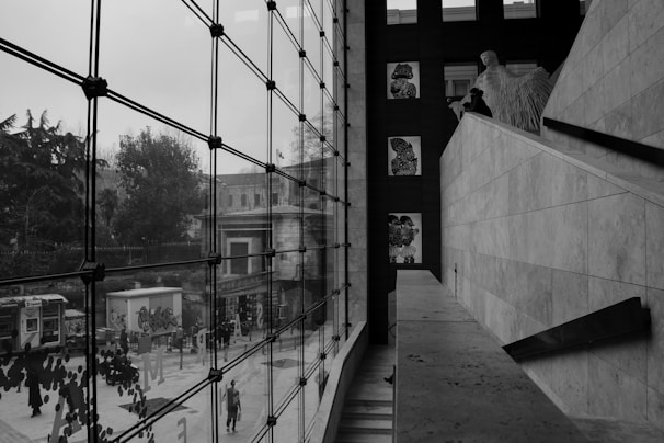 A modern interior space with large glass windows overlooking an urban street scene. The architecture features clean, geometric lines and art is displayed on a wall. Outside, there are trees, a phone booth, and people walking.