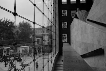 A modern interior space with large glass windows overlooking an urban street scene. The architecture features clean, geometric lines and art is displayed on a wall. Outside, there are trees, a phone booth, and people walking.