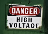 A warning sign labeled with the words 'DANGER HIGH VOLTAGE' on a metal plate. The background is painted a deep green, and the sign has a combination of red and black text on a white background. The corners of the metal plate are bolted, showing signs of wear and age.
