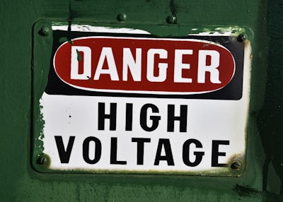 A warning sign labeled with the words 'DANGER HIGH VOLTAGE' on a metal plate. The background is painted a deep green, and the sign has a combination of red and black text on a white background. The corners of the metal plate are bolted, showing signs of wear and age.