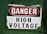 A warning sign labeled with the words 'DANGER HIGH VOLTAGE' on a metal plate. The background is painted a deep green, and the sign has a combination of red and black text on a white background. The corners of the metal plate are bolted, showing signs of wear and age.