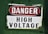 A warning sign labeled with the words 'DANGER HIGH VOLTAGE' on a metal plate. The background is painted a deep green, and the sign has a combination of red and black text on a white background. The corners of the metal plate are bolted, showing signs of wear and age.