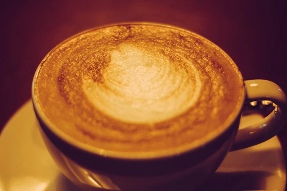 Close-up photo of a steaming cappuccino in an earthy ceramic cup with subtle mountain silhouette patterns in the background.