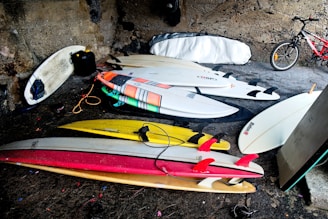 A collection of surfboards is neatly arranged on the ground in an enclosed area with a stone wall backdrop. The boards vary in color and design, including white, yellow, and red shades. A bicycle is parked nearby, partially visible against the wall. There is also a surfboard bag leaning against the wall and a black jerry can close to one of the boards.