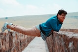 A person is wearing a denim jacket, checkered shirt, and beige pants while balancing sideways on a stone wall outdoors. The setting appears to be a rural, open area with grassy hills in the background.