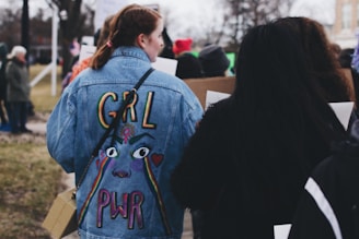 A group of people walking outdoors, with the focus on a person wearing a denim jacket featuring colorful embroidery with the words 'GRL PWR'. They appear to be part of a larger gathering or protest, as others around them hold signs.