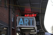 A building facade features a prominent sign with large blue letters spelling 'ART' and a red neon sign above it displaying 'GALLERY.' The architectural style is indicated by brick walls and arched windows. There is a secondary sign suggesting the presence of a bar and kitchen inside.