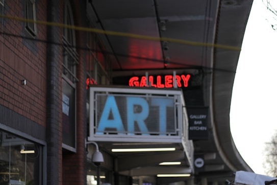 A building facade features a prominent sign with large blue letters spelling 'ART' and a red neon sign above it displaying 'GALLERY.' The architectural style is indicated by brick walls and arched windows. There is a secondary sign suggesting the presence of a bar and kitchen inside.