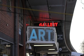A building facade features a prominent sign with large blue letters spelling 'ART' and a red neon sign above it displaying 'GALLERY.' The architectural style is indicated by brick walls and arched windows. There is a secondary sign suggesting the presence of a bar and kitchen inside.