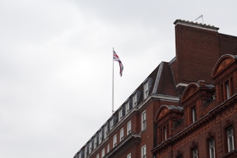 A large, red-brick building with classic architectural details. A Union Jack flag is mounted on a pole atop the building. The sky is overcast with a light gray color.