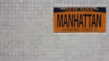An orange and black Manhattan license plate is mounted on a white tiled wall. The plate features the words 'New York' at the top, 'Manhattan' in the middle, and 'Empire State' at the bottom.
