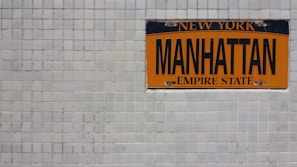 An orange and black Manhattan license plate is mounted on a white tiled wall. The plate features the words 'New York' at the top, 'Manhattan' in the middle, and 'Empire State' at the bottom.