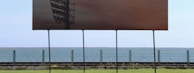 A large billboard with a dark image stands on a green grassy area, with the ocean and a clear sky in the background. The billboard displays a shadowy structure that resembles an industrial framework or tower. A metal fence separates the grassy area from the water along the horizon.