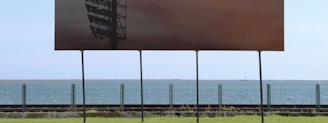 A large billboard with a dark image stands on a green grassy area, with the ocean and a clear sky in the background. The billboard displays a shadowy structure that resembles an industrial framework or tower. A metal fence separates the grassy area from the water along the horizon.
