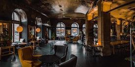 A stylish, dimly-lit caf&eacute; or lounge with arched windows and a mixture of vintage and modern d&eacute;cor. There are several seating areas with mismatched furniture including armchairs and small round tables. Warm ambient lighting from spherical lamps adds a cozy atmosphere. Plants are decoratively placed throughout the space.