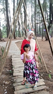 A woman and a child are standing on a wooden pathway in a forested area with tall pine trees surrounding them. The woman is wearing a red top and a light headscarf, and the child is in a pink and black dress with colorful flip-flops. The pathway is made of wooden planks and there are bamboo poles forming a triangular structure overhead.