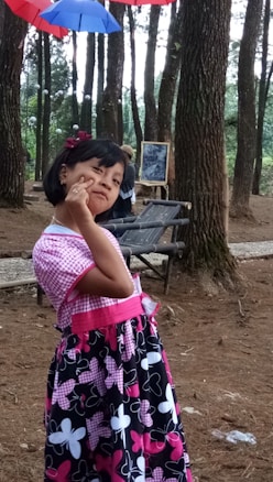 A young girl wearing a colorful dress stands playfully in a wooded area. Her dress features pink and black with large floral patterns. Above her, bright umbrellas in red and blue are hung, adding a whimsical touch to the forest setting. In the background, there is a framed picture and a wooden bench.