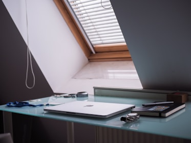 A minimalistic workspace featuring a closed laptop on a glass desk next to a window with blinds. Additional items include a notebook and pen, a wristwatch, glasses, a container, and some loose cables.