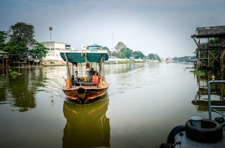 A small wooden boat with a covered canopy navigates a calm, reflective river. The boat carries several passengers and is adorned with colorful decorations. On the riverbanks, there are a mix of trees, residential buildings, and a wooden stilted house. The atmosphere is serene, with a clear, bright sky above.