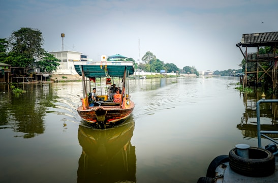 A small wooden boat with a covered canopy navigates a calm, reflective river. The boat carries several passengers and is adorned with colorful decorations. On the riverbanks, there are a mix of trees, residential buildings, and a wooden stilted house. The atmosphere is serene, with a clear, bright sky above.