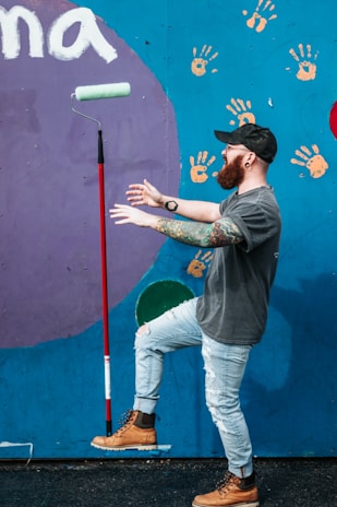 A man is playfully balancing a paint roller on its end while posing in front of a colorful mural. The wall behind him is decorated with handprints and a large circular shape. He is wearing a cap, glasses, and has tattoos on his arm, dressed in a t-shirt, ripped jeans, and boots.