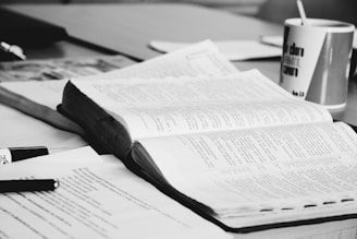 An open book with visible text lies on a table surrounded by other papers and a pen. A mug with text is also present, suggesting a study or work environment.