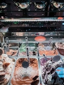 A display of various flavors of gelato in a shop, with each tray featuring a distinct flavor labeled with signs written in multiple languages, including Hebrew. The ice cream includes flavors like chocolate, mint, and berry, with decorative swirls and toppings. Reflective metal trays and serving scoops are arranged neatly within a glass-covered counter.