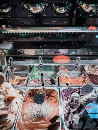A display of various flavors of gelato in a shop, with each tray featuring a distinct flavor labeled with signs written in multiple languages, including Hebrew. The ice cream includes flavors like chocolate, mint, and berry, with decorative swirls and toppings. Reflective metal trays and serving scoops are arranged neatly within a glass-covered counter.