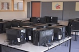 A computer lab with several rows of black desktop computers placed on tables, each connected with various cables. The room has a blue carpet and the walls have bulletin boards with posters. The arrangement suggests a classroom or training environment.