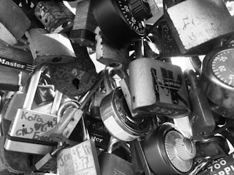 A collection of different padlocks and combination locks clustered together, with some locks displaying inscriptions or engravings. The locks vary in shape and design, creating an intricate pattern resembling a makeshift sculpture.