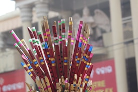 A cluster of colorful wooden flutes with vibrant paint and intricate patterns, arranged standing up. In the background, a blurred building with columns and what appears to be a statue is visible.