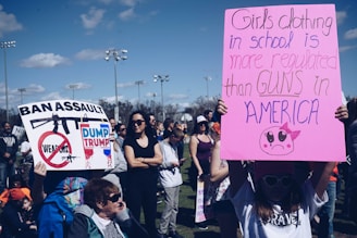 A group of people gathered outdoors, many holding protest signs. One prominent sign on the left reads 'Ban Assault Weapons' with a graphic of a gun crossed out, and another sign says 'Dump Trump'. Another large pink sign on the right has a message about the regulation of girls' clothing versus guns in America. The event appears to be a public protest or rally, with individuals of various ages present.