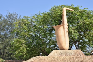 A large sculpture of a saxophone made from wicker material is prominently displayed against a backdrop of lush green trees. The saxophone stands atop a structure covered in straw, and clear blue skies are visible above.