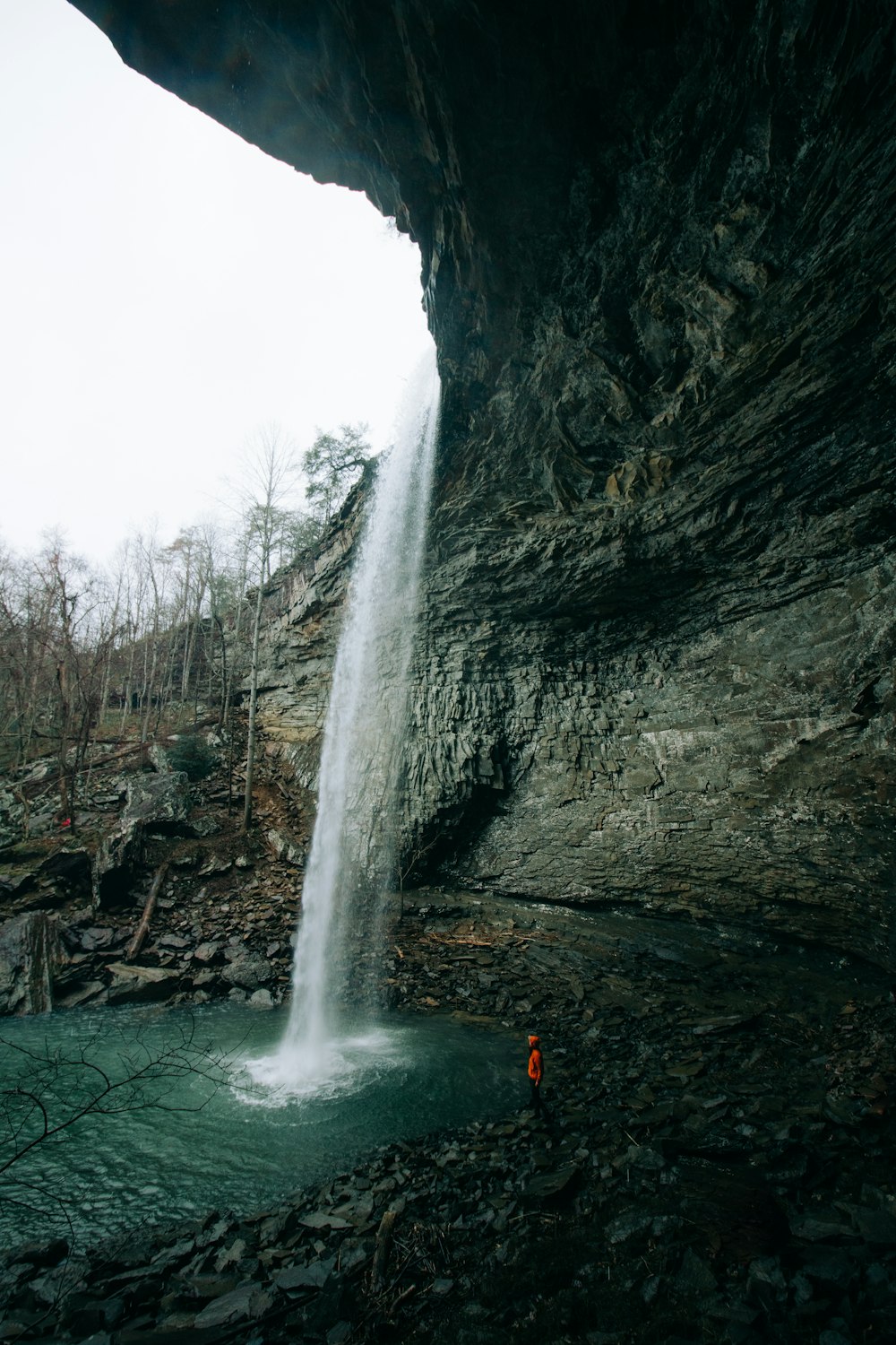 man standing under cave waterfall under cloudy skies